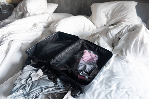 Open suitcase with clothing on messy white bed in hotel. - 写真・画像