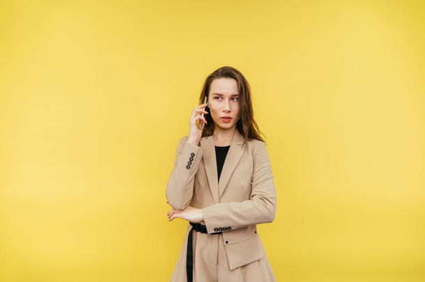 Attractive business woman in a jacket talks on the phone on a yellow background with a serious face and looks away. - Foto, afbeelding
