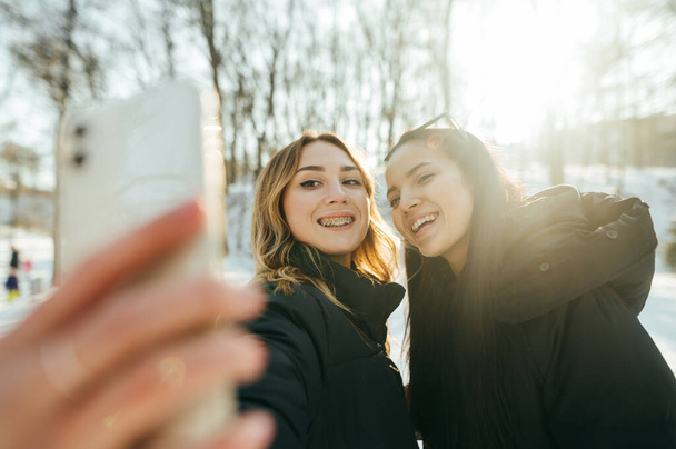 İki güzel mutlu kız arkadaş kış ormanında yüzlerinde tebessümle akıllı bir telefondan selfie çekiyor. En iyi arkadaşlar parkta yürürken fotoğraf çekilirler.. - Fotoğraf, Görsel