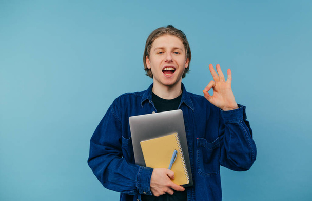Πορτρέτο ενός θετικού μαθητή σε πουκάμισο στέκεται σε μπλε φόντο με ένα φορητό υπολογιστή και σημειωματάρια στα χέρια του και κοιτάζοντας την κάμερα με ένα χαμόγελο στο πρόσωπό του, δείχνει μια χειρονομία του ok - Φωτογραφία, εικόνα