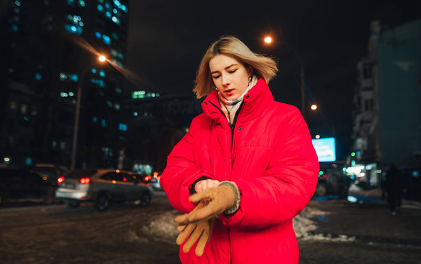 Woman in a jacket stands in the winter night in the cool temperature on the street and puts on mittens on his hands on the background of the night city. - Photo, Image