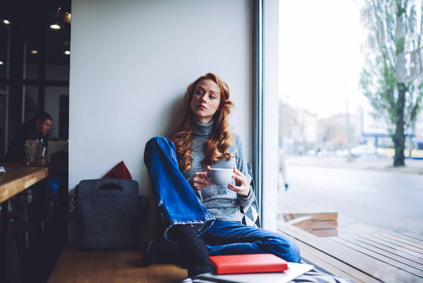 Young pensive long haired female in casual outfit sitting with crossed legs on window sill drinking hot beverage and looking away in cafe - Zdjęcie, obraz