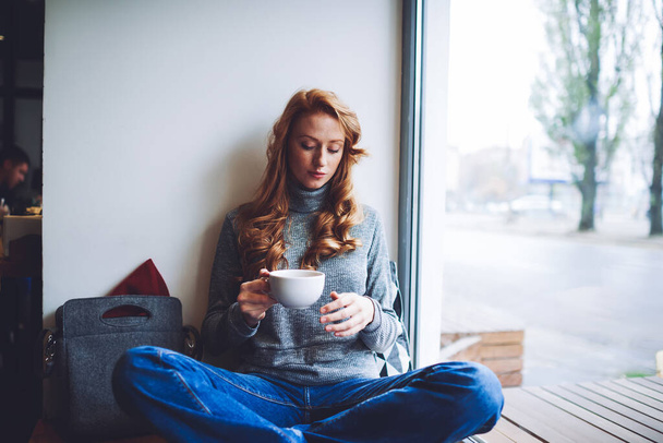 Pensive female in casual clothes sitting near window with crossed legs and closed eyes drinking coffee while having rest in light cafe - Foto, afbeelding