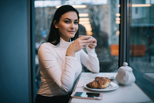 Satisfied female in casual outfit with long hair looking at camera while drinking coffee having rest in light modern cafeteria - Foto, Bild