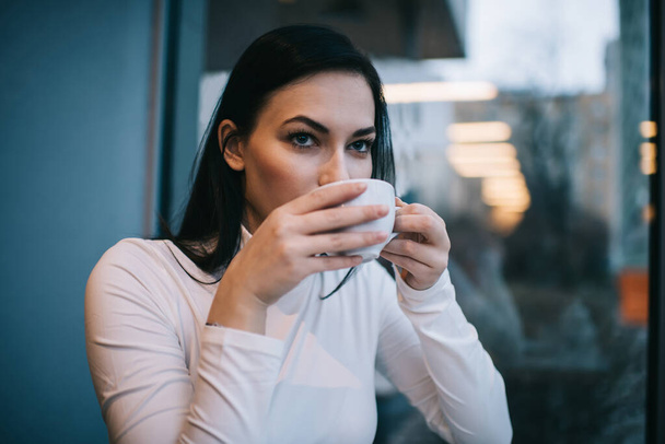 Thoughtful female in white clothes with dark hair looking away while drinking coffee form white cup resting in light cafe - Photo, Image