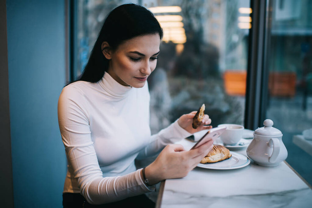 Focused female with long hair sitting at table in cafe and using mobile phone for text messaging while eating croissant during break - 写真・画像