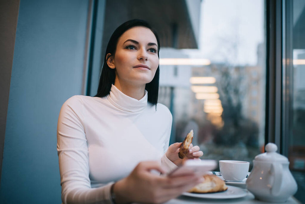 Glad woman wearing casual outfit with long hair looking away and text messaging on mobile phone while eating croissant during break - 写真・画像