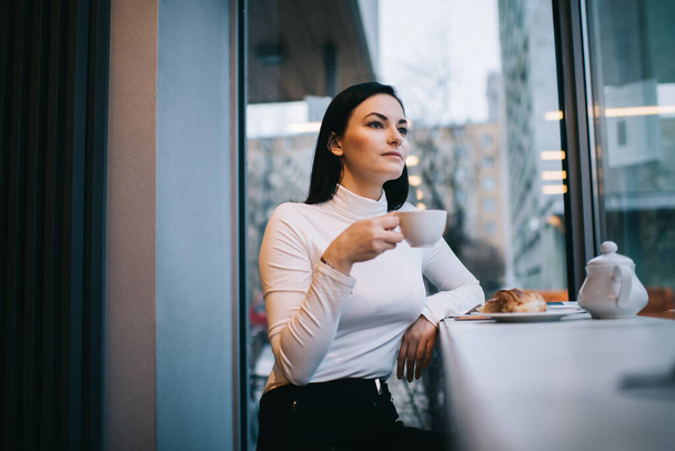 Thoughtful female with long dark hair looking away and sitting with white cup of hot coffee and croissant in cafeteria - Photo, Image