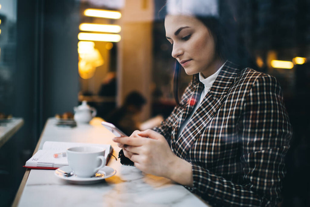 Side view of concentrated female in stylish clothes sitting at table in cafe with cup of coffee and messaging on smartphone - Foto, immagini