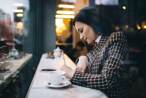 Side view of pensive female student sitting at table with cup of coffee and taking notes while doing task for university - 写真・画像