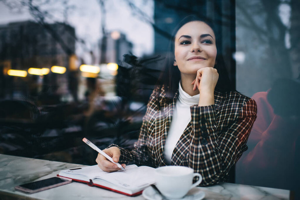 Through window on satisfied female remote worker in stylish checkered jacket looking away while sitting at table with planner and working - Фото, изображение