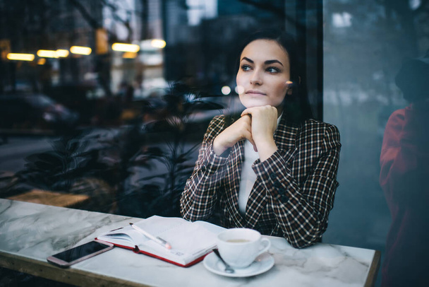 Through glass window of thoughtful female entrepreneur in stylish clothes sitting at table with cup of beverage and opened notebook and pensively looking away in daytime - Foto, Imagen
