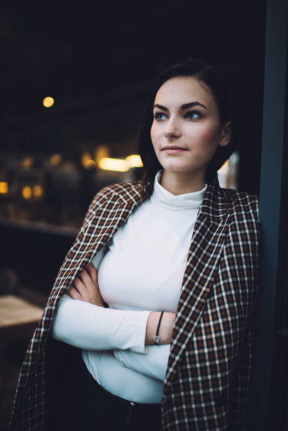 Young confident female with dark hair in trendy outfit looking away while leaning on wall with crossed arms in modern cafe - Фото, изображение