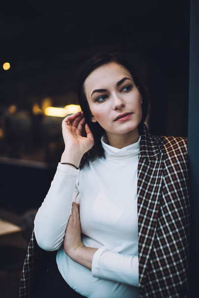Young stylish female with black hair in elegant clothes looking away thoughtfully while standing against wall and touching hair in modern cafeteria - Fotoğraf, Görsel