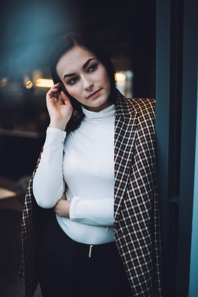 Young focused black haired female in stylish outfit standing against wall while looking away thoughtfully and touching hair in modern cafe - Foto, afbeelding