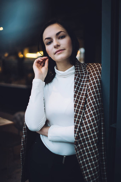 Confident young lady wearing plaid coat and turtleneck standing near black wall in modern cafe touching hair and looking at camera - Photo, Image