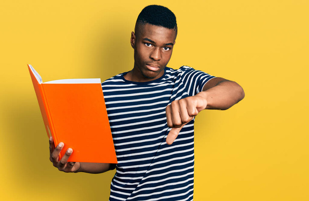 Jeune homme afro-américain tenant le livre avec le visage en colère, signe négatif montrant aversion avec les pouces vers le bas, concept de rejet  - Photo, image