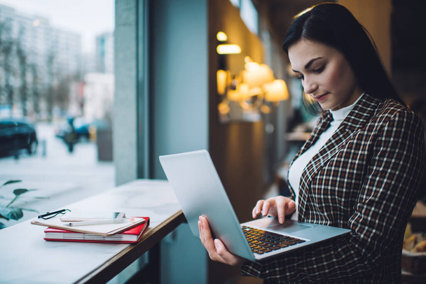 Young female freelancer in elegant clothes working on computer while standing in cafe near notebook and smartphone near clipboard on table in daytime - 写真・画像