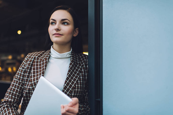 Confident young woman in elegant outfit leaning on wall while standing with computer in hands in cafe entrance in day and looking away - 写真・画像