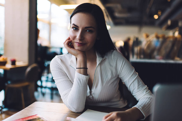 Smiling female wearing casual outfit looking at camera leaning on hand while sitting at table on blurred background of modern cafe - Foto, Imagem