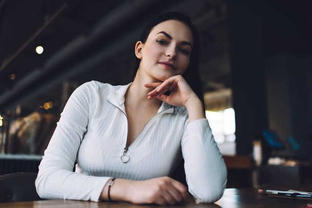 Serious female in white casual outfit looking at camera while sitting at table in cafeteria with hand at chin during break - Фото, изображение