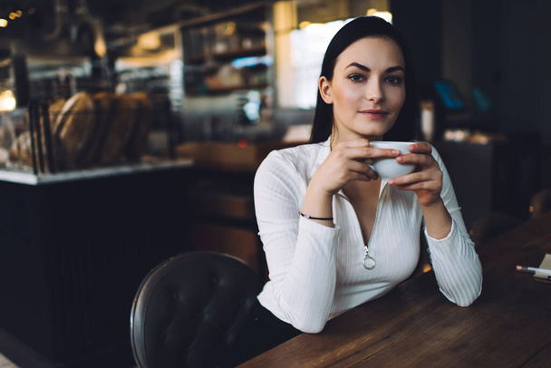 Emotionless young female in white blouse with long dark hair drinking coffee sitting at table in cafe and looking at camera - Fotografie, Obrázek