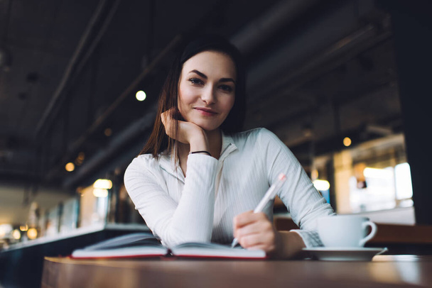 Positive young brunette female student in trendy clothes sitting at table with cup of coffee and smiling while taking notes in diary in contemporary cafe - Foto, Bild