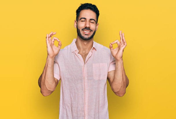Homem hispânico com barba vestindo camisa casual relaxar e sorrir com os olhos fechados fazendo gesto de meditação com os dedos. conceito de ioga.  - Foto, Imagem