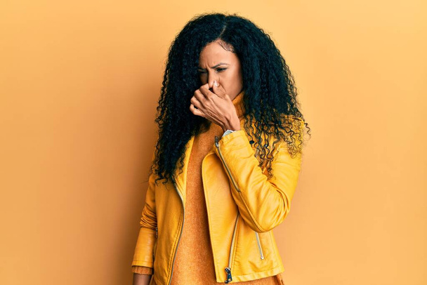 Középkorú afro-amerikai nő gyapjú téli pulóvert és bőrdzsekit visel, bűzlik valami bűzös és undorító, elviselhetetlen szagtól, visszatartja a lélegzetét ujjakkal az orrán. rossz szag.  - Fotó, kép