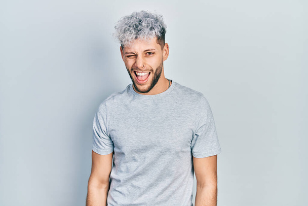 Jeune homme hispanique aux cheveux teints modernes portant un t-shirt gris décontracté clignant des yeux en regardant la caméra avec une expression sexy, un visage joyeux et heureux.  - Photo, image