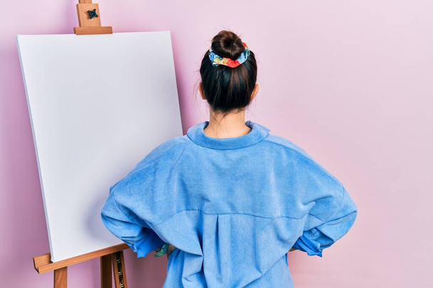 Young hispanic woman standing by painter easel stand standing backwards looking away with arms on body  - Photo, image