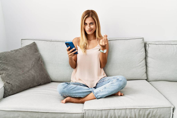 Blonde beautiful young woman sitting on the sofa at home using smartphone doing money gesture with hands, asking for salary payment, millionaire business  - Valokuva, kuva