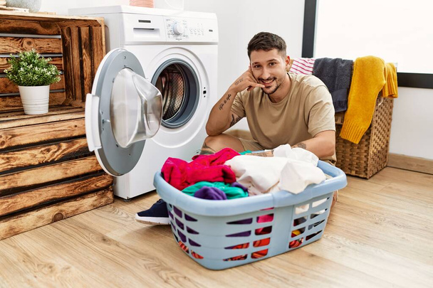 Young handsome man putting dirty laundry into washing machine smiling doing phone gesture with hand and fingers like talking on the telephone. communicating concepts.  - Foto, Imagem