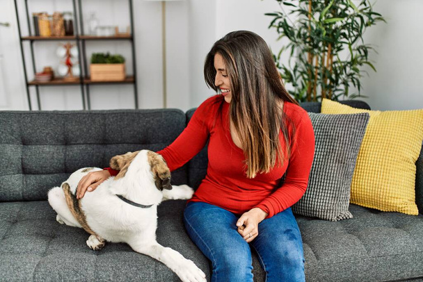 Молода жінка посміхається впевнено сидить на дивані з собакою вдома
 - Фото, зображення