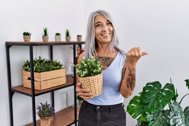 Mujer de pelo gris de mediana edad sosteniendo maceta verde en casa apuntando hacia atrás con la mano y los pulgares hacia arriba, sonriendo confiada  - Foto, Imagen