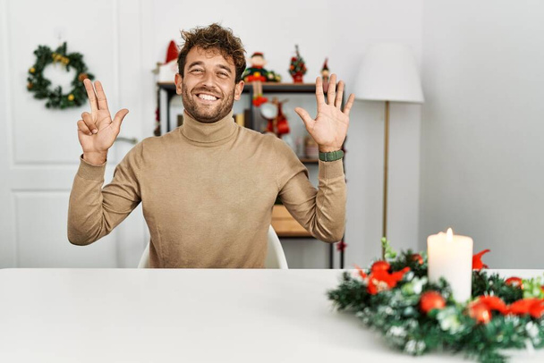 Young handsome man with beard sitting on the table by christmas decoration showing and pointing up with fingers number eight while smiling confident and happy.  - Fotó, kép