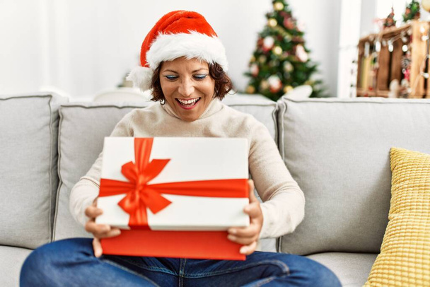 Hispanische Frau mittleren Alters entpackt Geschenk auf dem Sofa am Weihnachtsbaum zu Hause. - Foto, Bild