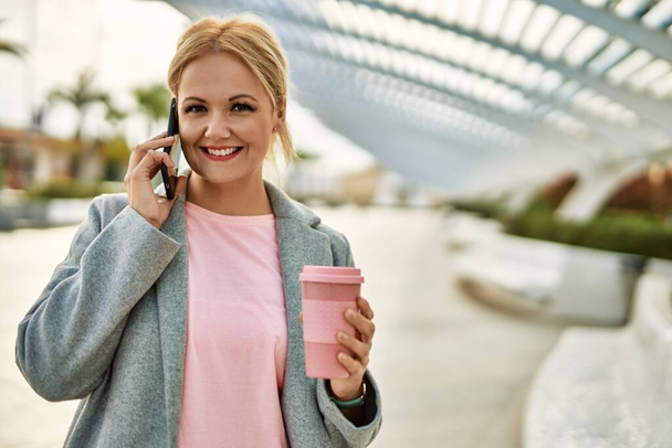 Joven mujer de negocios rubia hablando en el teléfono inteligente tomando café en la ciudad. - Foto, imagen