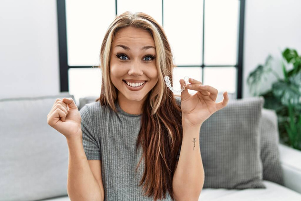 Young caucasian woman holding invisible aligner orthodontic screaming proud, celebrating victory and success very excited with raised arm  - Foto, imagen