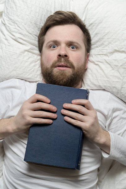 Tyrmistynyt mies makaa sängyssä kiinni kirjan hän on valmis rinnassa ja etsii innokkaasti. Yläkuva.  - Valokuva, kuva