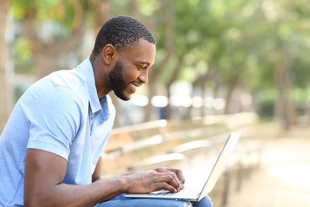 Side view portrait of a happy man with black skin using laptop sitting on a bench in a park - Fotó, kép