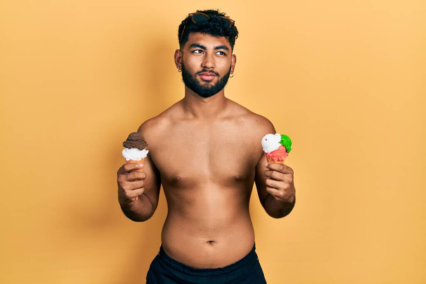 2つのアイスクリームコーンを食べて泳いで身に着けている髭を生やしているアラブ人男性側を見て笑って離れて考えて見つめて.  - 写真・画像