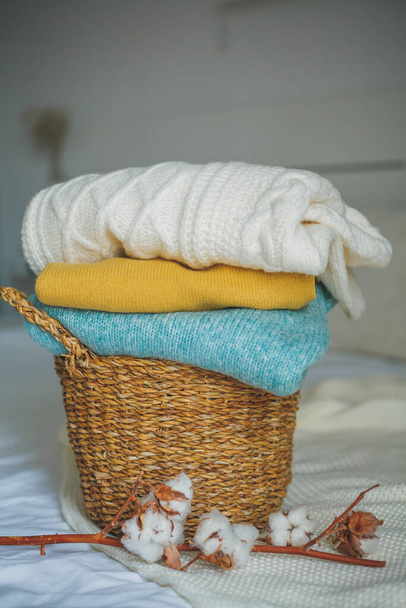 Stack of knitted warm sweaters in wicker rattan basket, plaid, branch of cotton. Autumn mood. Fall. - Photo, image