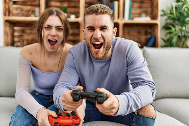 Νεαρό καυκάσιο ζευγάρι ουρλιάζοντας και παίζοντας βιντεοπαιχνίδι στο σπίτι. - Φωτογραφία, εικόνα