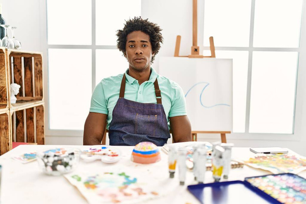 Jovem afro-americano sentado em cima da mesa no estúdio de arte com expressão séria no rosto. simples e natural olhando para a câmera.  - Foto, Imagem