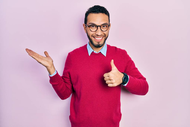 Hispánec s vousy v obchodní košili a brýlích ukazující dlaň a dělá v pořádku gesto s palci nahoru, usmívá šťastný a veselý  - Fotografie, Obrázek