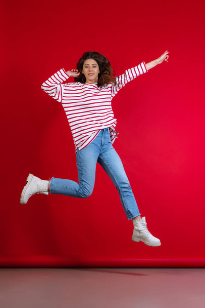 Emotional young beautiful girl in sweater and jeans jumping isolated on red studio background. Concept of emotions, facial expression - Foto, Imagen
