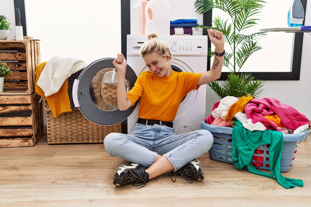 Nuori blondi nainen tekee pyykkiä istuu pesukone tanssia onnellinen ja iloinen, hymyillen liikkuvat rento ja luottavainen musiikin kuunteluun  - Valokuva, kuva