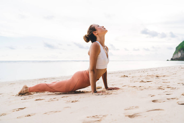 Side view of unrecognizable flexible female in sporty costume practicing high cobra position while training asana on sand near sea - Foto, imagen