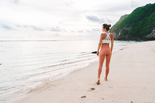 Back view of unrecognizable female in stylish sportswear standing on sandy beach near waving sea and enjoying picturesque seascape against rocky cliff with green plants in sunny day - Foto, Imagem